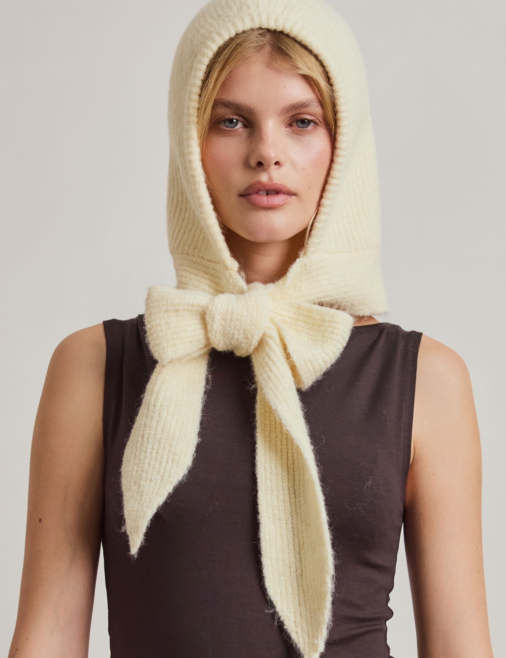 A model wearing a cream-colored fuzzy knit headscarf with a large bow, styled over a dark brown sleeveless top and beige trousers, against a plain light gray studio background.