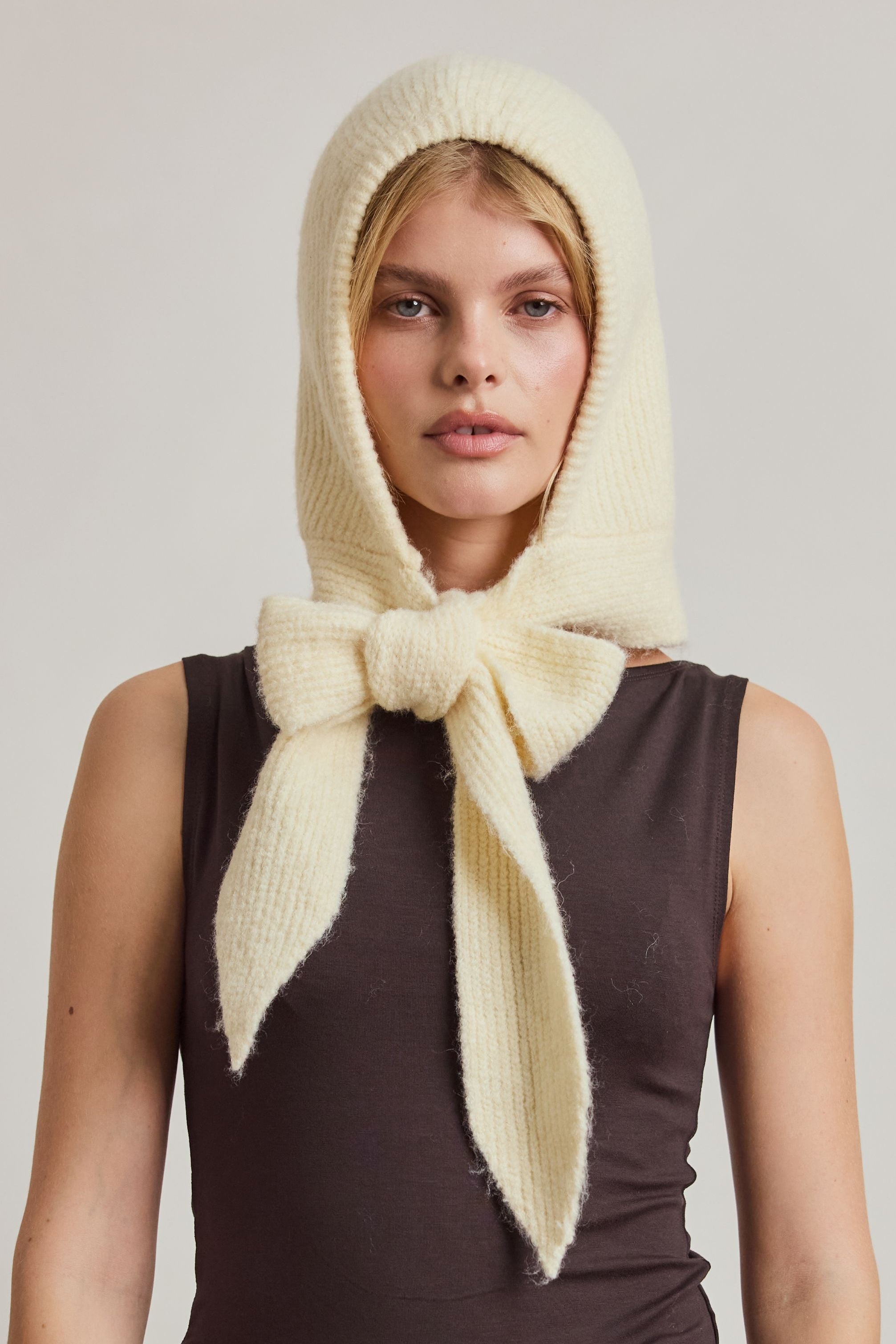 A model wearing a cream-colored fuzzy knit headscarf with a large bow, styled over a dark brown sleeveless top and beige trousers, against a plain light gray studio background.